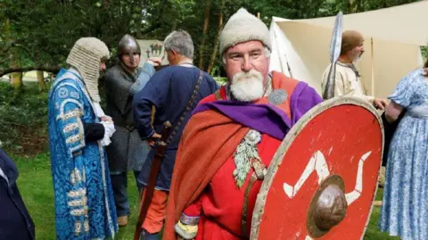 Martin Parr/Magnum Photos A man dress as a Viking holds a red shield with with a triskelion. Others in traditional dress, including a man in a wig, stand around behind him.