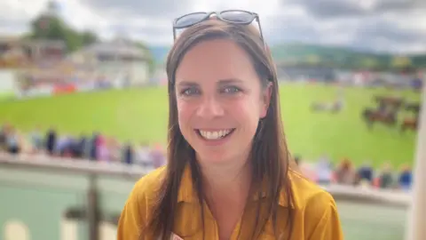 Teleri Fielden, policy officer for the Farmers' Union of Wales stands in front of the main ring at the Royal Welsh Agricultural Show.  She is has long brown hair, is wearing a yellow shirt.