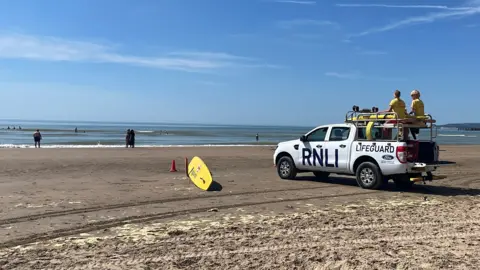 George Carden/BBC A white RNLI van on a sand beach with two lifeguards on top.