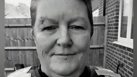 Sharon OFogarty A black and white image of Sharon OFogarty. He has eyebrow and lip piercings. She is standing outside next to a building with a fence in the background. She is wearing a security uniform. 