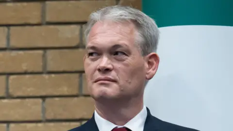 A man with white hair and wearing a suit has a blank expression on his face