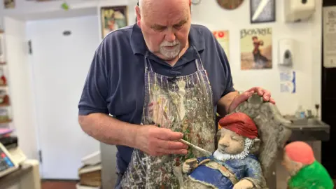 Alex Dunlop/BBC Alistair Dent with a paint brush in hand working on a gnome. The gnome is slumped in a chair and is wearing a blue outfit with a red hat. Dent is wearing a blue short-sleeved navy polo shirt with a blue and white striped apron covered in paint over the top.