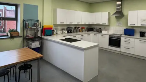 The Way The kitchen has light green walls, white cupboards and a table on the left.