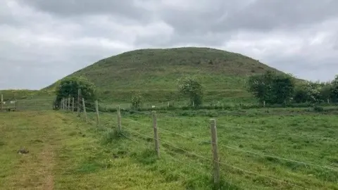 A large mound of earth covered in grass, including a grass field in the foreground with a fence running alongside a footpath. 