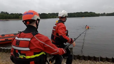 Two firefighters stand on the side of a lake and use a long black pole to help rescue a casualty in the water