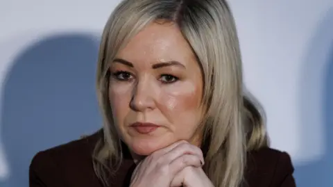 A close-up of Michelle O'Neill during a press conference at the British-Irish Council in Wales in December 2025. She is a woman with straight, shoulder-length blonde hair. She is wearing a dark suit jacket and is resting her face on her hands, as though listening to someone speak.