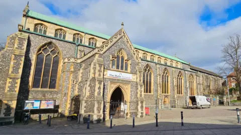 We can see the outside of St Andrews Hall on a sunny day. It is a medieval building, but there is some fencing and a works van outside of it.