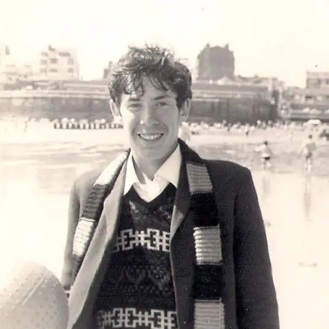 Richard Dowell A black and white picture of a young man on Bridlington beach. He has a jacket on over a knitted pullover and has a scarf around his neck.