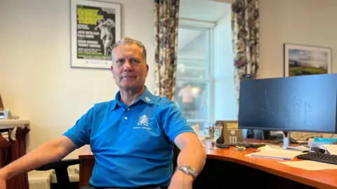Darren Attwood looks at the camera as he sits in a chair with his arms on the arms of the chair and his desk is next to him. He's wearing a blue t-shirt and has grey slicked-back hair.