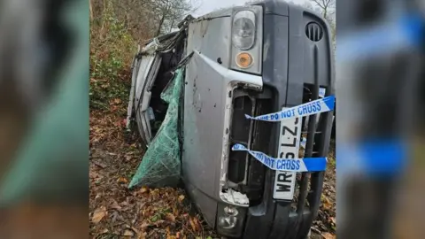 Tyler O'Brien A damaged silver van lays on its side with a smashed, folded windscreen. It is among woodland. There is police tape wrapped around its bonnet.