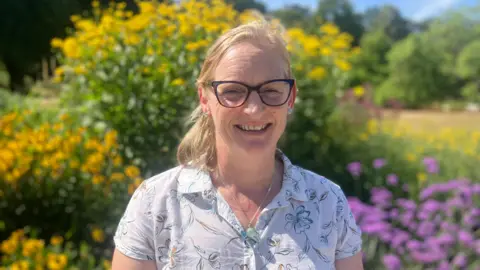 Elizabeth Baines / BBC A woman wears a white shirt and glasses. She has blonde hair and stands in front of a herbacious border which is blurred behind.