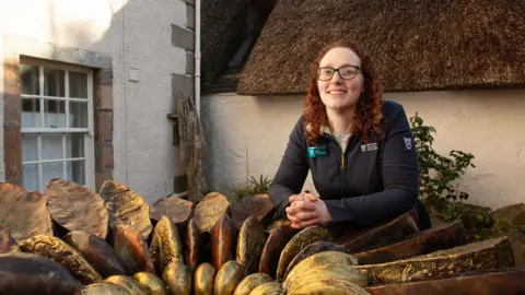 National Trust for Scotland Debbie Reid has long curly auburn hair and is wearing dark-rimmed glasses and a National Trust for Scotland zipped top. She is leaning on a sculpture of an ammonite outside the Hugh Miller's Birthplace Cottage and Museum in Cromarty.