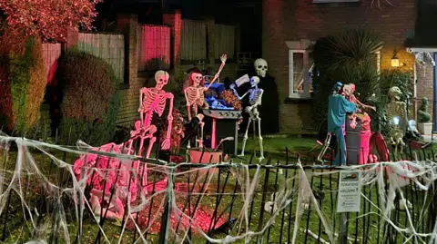 Kerry Morgan A house lit by red lights with skeletons decorating the garden in various poses including around a barbecue. A fence in the foreground is decorated with fake cobwebs