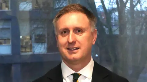 John Fairhall/BBC Ed Garrett smiles at the camera in front of a large window. He wears a black suit with a green and yellow tie. He has short grey hair.