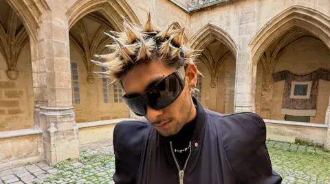 Aly Meghani is looking down, wearing huge black sunglasses and a black jacket. His hair is in blonde spikes.