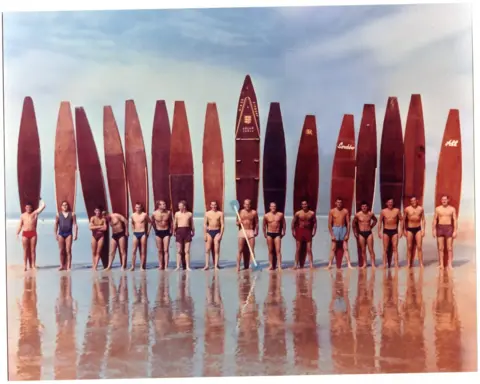 John Houillebecq A photo which shows 16 people stood in a line on a beach. Behind each of them is a surfboard. In the background is blue sky with white clouds. 