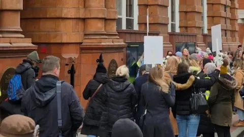 Protesters gather on a street with placards and signs outside the red bricked entrance to the Greater Manchester Combined Authority's Oxford Street office.