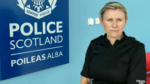 Ch Supt Emma Croft wears a black police uniform and sits in front of a light blue wall next to the dark blue Police Scotland crest