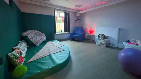 Staffordshire County Council A sensory room in a care centre. The walls are green and white. A thick, curved green mat is in the corner with cushions on it. A blue armchair is visible in the corner and there is a purple medicine ball on the right.