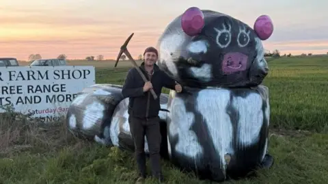 Newborough YFC A man leaning against a model bull made of round straw bales covered in black plastic painted with white markings. It has a pink painted nose with a ring in it and pink ears. The man has a pickaxe over his shoulder, is smiling and has his left thumb up. Behind him are flat arable fields and a sunset. 