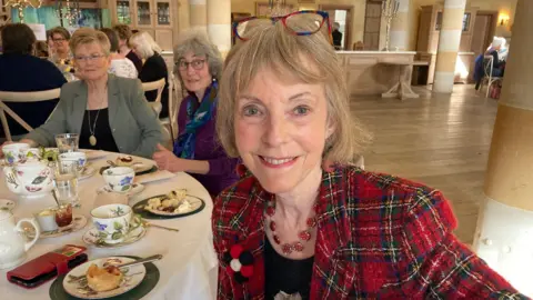 Una mujer de cabello rubio canoso sonríe y mira a la cámara sentada alrededor de una mesa disfrutando de un té con otras mujeres. Lleva una chaqueta de tartán roja y un collar rojo. Lleva gafas de carey marrones y azules.