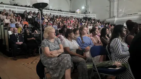 Sam Read/BBC People sit in a large hall in the Bedford Corn Exchange in rows and are looking towards a stage (not in image).