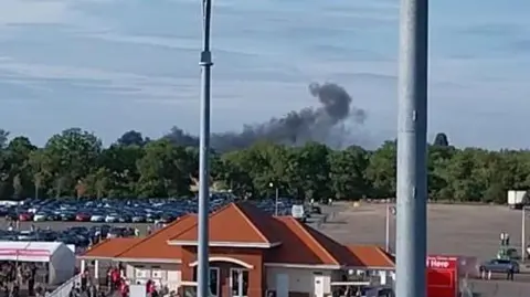 Black smoke rising above trees into the sky at Chelmsford City Racecourse. In front of the trees is a large car park with many cars. In front of them is a low-lying red roofed building. People can be seen walking up to a tented entrance to the venue. 