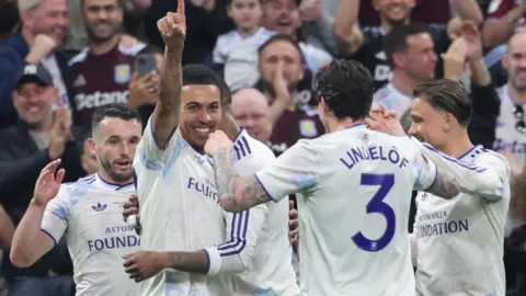 Several Villa players gather round Rogers who has his right hand in the air after scoring. They have a white kit on with blue numbers on their backs.