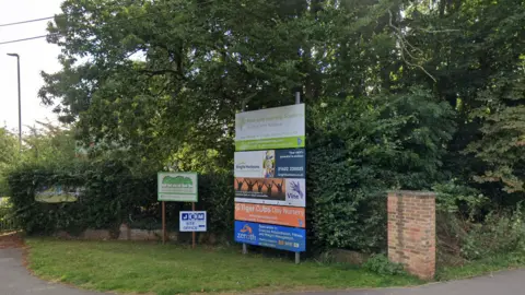 Signage on a grass verge advertising New Line Learning Academy, in green, ans a few other schools. The other signs are blue, orange, and navy and white.