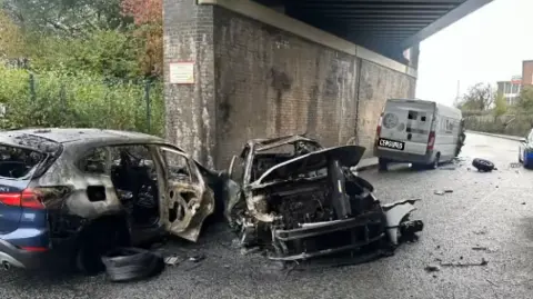 Bilston Fire Station Two burnt out cars sit underneath a railway bridge. Both have suffered extensive damage and look completely destroyed. The van is parked a few feet away and has suffered fire damage, although it is less severe.