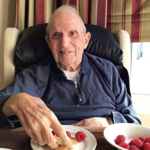 BBC Elderly man smiling while sitting at a table, reaching for food on a plate with raspberries, with striped curtains in the background.
