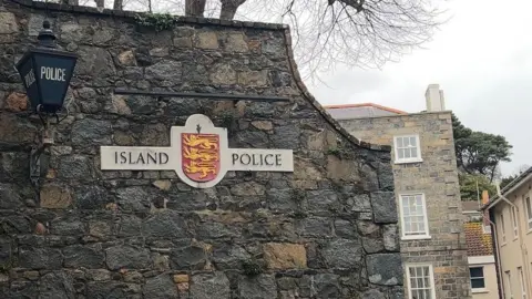 BBC A photo of a granite wall with a sign that says 'Island Police' on with with a red badge with 3 golden lions in the middle.