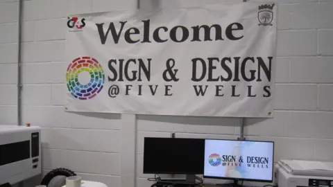 Alex Pope/BBC A sign and design room, in a prison with two computer screens, one is on. The walls are painted white. 