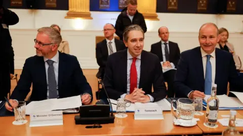 EPA Taoiseach and Fine Gael leader Simon Harris (middle) , Fianna Fáil leader Micheál Martin (right) and Green Party leader Roderic O'Gorman (left) sit at a table with name tags in front of them. Simon Harris is wearing a bright red tie and dark blazer, Roderic is wearing a black and white speckled tie and wears glasses. Micheál is wearing a light blue tie and navy blazer.