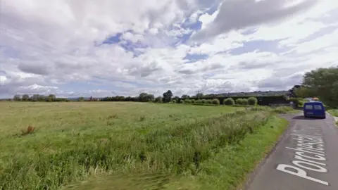 StreetView image of the land earmarked for development. There is a single-track road on the right of the picture, and to the left an open green field with power lines running over it. 