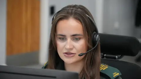 Shaun Whitmore/BBC Nicole Douglas has long brown hair and she is wearing a green NHS call handler uniform. She is sitting on a chair in an office and is looking at a computer wearing a black headset.