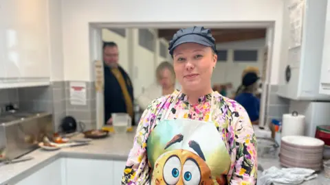 Maddie Riley stands in the kitchen at Water Orton's Methodist Church, behind her people are queuing up for jacket potatoes. She wears a floral blouse underneath an apron featuring a cartoon potato. She has a catering cap and her hair tied up.