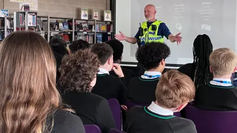 Sam Read/BBC A class of children seen from behind listening to a police officer speak about e-scooters. He has a PowerPoint presentation behind him.