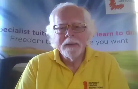 John Rogers, Chief Executive of the Association of Disability Driving Instructors, He is wearing a yellow polo shirt and has glasses, grey hair and a grey goatee.