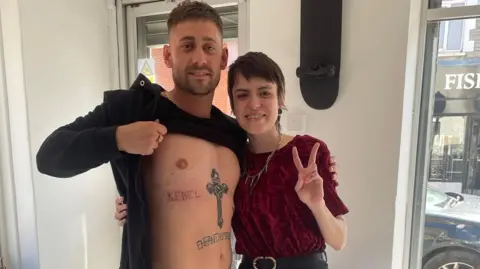 Bianca Oliveira Bianca Oliveira and Michael Socha. Bianca holds up her fingers in a piece sign. She wears a red velvet top and has short brown hair. Michael (left) holds up his t shirt to show tattoo which reads: rebel. He has brown hair and a brown short beard.