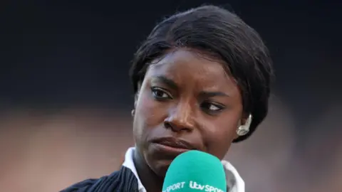 Getty Images Eni Aluko looks quizzically as she works on ITV's football coverage in 2024, while holding a turquoise ITV Sport microphone. She has black hair tied back and wears a broadcast earpiece.