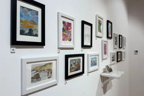A picture at an angle showing 13 frames with art in them . There is a small shelf in the middle of the display with a ceramic piece.