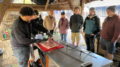 Adam Purnell is on the left wearing gloves, with one glove holding meat that is on a table outside. Six other people are in the photo, with most of them on the opposite side of the table to Purnell.