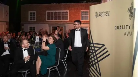 Men and women in black tie and evening dresses are sitting at tables, applauding as the awards are announced at a ceremony. A big poster with "West Mercia Police" on it sits to one side.