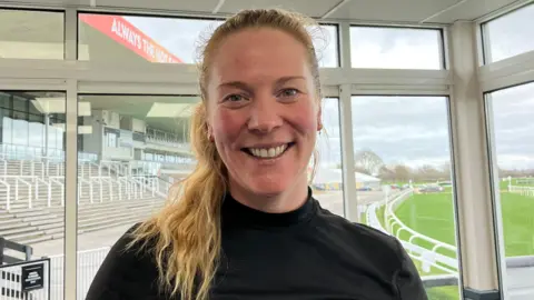 A woman with long blonde hair in a pony tail, draped over her right shoulder, and wearing a black top, smiles as she stands in a room with windows behind her. A race track with stands next to it can be seen outside.