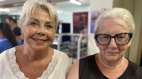 A picture of two women. Jenny Scott-Jones is wearing a necklace and a white top and has short, white hair. Iris Anderson also has short, white hair. She is wearing dark-rimmed glasses, a white necklace and a black top.