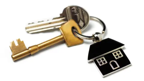 A set of house keys with a black house-shaped fob. One of the keys is long and gold in colour and the other is a smaller silver key. The set has been photographed against a white background.