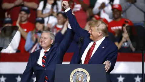 President Donald Trump and Senator Lindsey Graham are wearing navy blue business suits and standing behind a podium with the US presidential seal holding, each holding an arm up toward the other in a victorious gesture