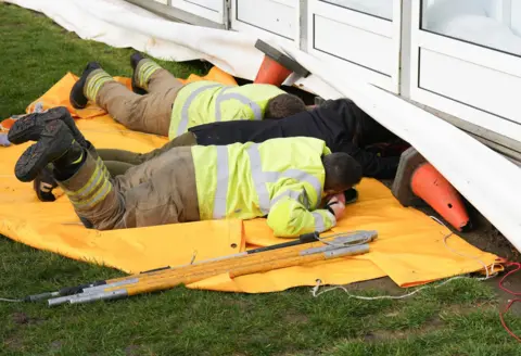 Eddie Mitchell Two firefighters and a woman looking underneath an ice rink. They are lying on top of a large orange tarpaulin with rescue tools on the side.