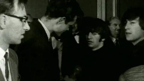 Ringo Star and John Lennon meet the press at the venue, then known as the ABC, when they performed there. This black-and-white image comes from BBC video footage. It shows Ringo in the centre of the image with distinctive sideburns. Jon Lennon is to his right.